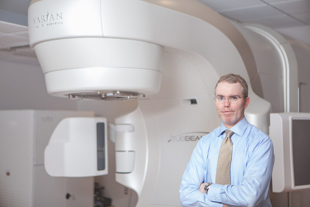 medium longshot of Dr. Jeffrey Brabham in blue shirt and gray tie standing in front of a Varian radiation machine.